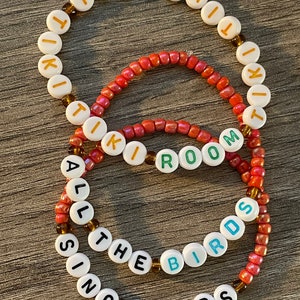 May include: Three beaded bracelets with white beads and brown spacers. The top bracelet spells "TIKI" in gold letters. The middle bracelet spells "ROOM" in green letters. The bottom bracelet spells "ALL THE BIRDS SING WORDS" in black letters.