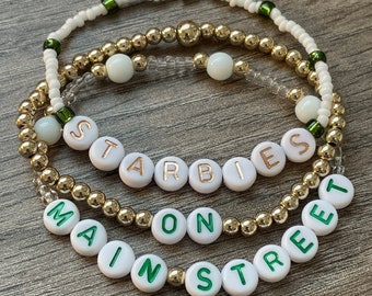 May include: Three white beaded bracelets with gold beads and green accents. The bracelets spell out "Starbies on Main Street" in white letters with gold outlines.