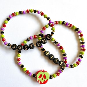 May include: Three beaded bracelets with the text "JUST ONE BITE". The bracelets feature a mix of red, green, purple, black, and gold beads. One bracelet has a red apple charm with a green skull design.
