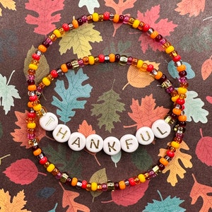 May include: A beaded bracelet with the word "THANKFUL" spelled out in white beads with gold lettering. The bracelet is made with orange, yellow, red, and purple beads.
