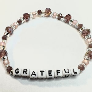 May include: A beaded bracelet with brown and pink beads and a white charm with the word "GRATEFUL" spelled out in black letters.