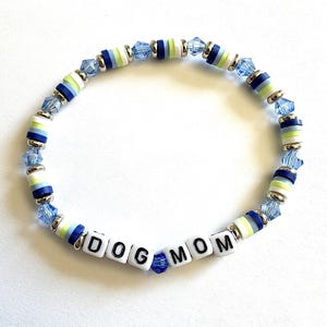 May include: A beaded bracelet featuring white, lime green, and dark blue polymer clay discs, interspersed with faceted light blue crystal beads and silver-toned spacers. The bracelet includes white square beads with black letters spelling DOG MOM.