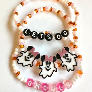 May include: Two beaded bracelets featuring white, orange, and pink beads. One bracelet includes black letter beads spelling LETS GO, while the other features three white ghost charms with black ears and pink bows, plus pink letter beads spelling GHOULS.