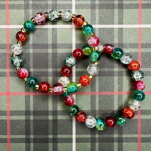 May include: Two bracelets made of colorful glass beads with a sparkly finish. The beads are red, green, white, and clear. The bracelets are on a plaid background.