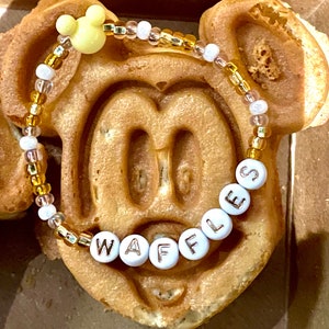 May include: A yellow Mickey Mouse head charm on a beaded bracelet with white and gold beads. The bracelet spells out "Waffles" in white letters.
