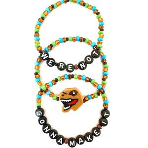 May include: Three beaded bracelets with the phrases "WE'RE NOT" and "GONNA MAKE IT". The bracelets feature a variety of colorful beads and a brown dinosaur head charm. The bracelets are on a white background.