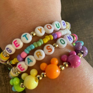 May include: A colorful beaded bracelet with the words "GET YOUR EARSON" spelled out in white letters on white beads. The bracelet also features Mickey Mouse head charms in yellow, orange, and purple.