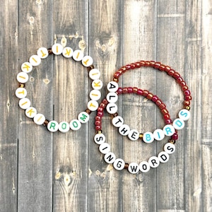 May include: Three beaded bracelets featuring white circular beads with black, yellow, and green lettering. One bracelet spells TIKI TIKI ROOM, while the other two combine to spell ALL THE BIRDS SING WORDS. The beads are strung on a thin, flexible cord.