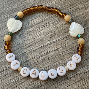 May include: A white beaded bracelet with brown and beige beads. The bracelet spells out "IT CALLS ME" in gold lettering on the white beads. The bracelet also has two white shell heart-shaped beads with a leaf design.