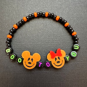 May include: A black and orange beaded bracelet with the words "BOO TO YOU" spelled out in green and purple letters. The bracelet also features two orange Mickey Mouse characters with black eyes and a red bow on the female character.