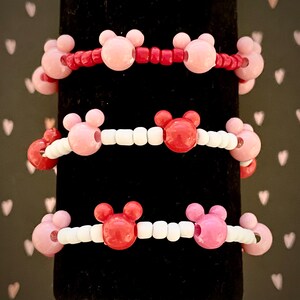May include: Three bracelets with pink and red Mickey Mouse head beads. The top bracelet has red beads, the middle bracelet has white beads, and the bottom bracelet has white beads.
