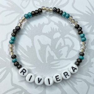 May include: A beaded bracelet with black, gold, and teal beads. The bracelet spells out the word "RIVIERA" in white letters on black beads.