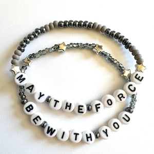 May include: Two beaded bracelets. The top bracelet is gray with silver beads and star charms. The bottom bracelet is white with black letters that spell "MAY THE FORCE BE WITH YOU".