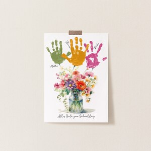 May include: A white card with three handprints in green, orange, and pink. The handprints are labeled with the names "Mattia", "Ben", and "Rika". A bouquet of flowers in a glass jar is below the handprints. The card says "Alles Gute zum Geburtstag" which is German for "Happy Birthday".