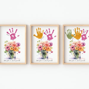 Op de afbeelding: Drie ingelijste prints met handafdrukken in verschillende kleuren, roze, oranje en groen, gerangschikt in een rij. Elke print toont een aquarel van een boeket bloemen in een glazen pot. De tekst "Mit Liebe von Hand gemacht" is onder elke afbeelding gedrukt.