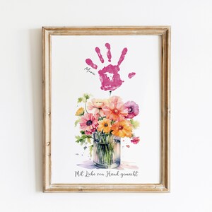 Op de afbeelding: Een ingelijst afdruk met een roze handafdruk en de naam "Maria" eronder. De handafdruk bevindt zich boven een aquarel van een boeket bloemen in een glazen vaas. De tekst "Mit Liebe von Hand gemacht" staat onderaan de afdruk.