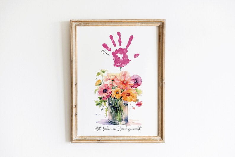 Op de afbeelding: Een ingelijst afdruk met een roze handafdruk en de naam "Maria" eronder. De handafdruk bevindt zich boven een aquarel van een boeket bloemen in een glazen vaas. De tekst "Mit Liebe von Hand gemacht" staat onderaan de afdruk.