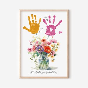 May include: A watercolor print with two handprints in yellow and pink, with the names "Mattea" and "Rike" written next to them. The handprints are above a bouquet of flowers in a glass jar. The text "Alles Gute zum Geburtstag" is at the bottom of the print.