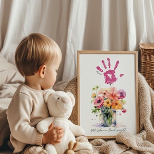 May include: A framed watercolor print of a bouquet of flowers in a glass vase. The flowers are pink, yellow, and orange. A pink handprint is above the flowers with the name "Maria" written below it. The text "Mit Liebe von Hand gemacht" is at the bottom of the print.