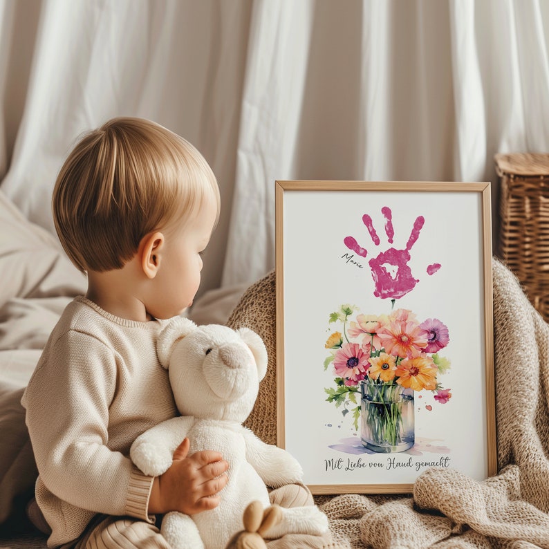 May include: A framed watercolor print of a bouquet of flowers in a glass vase. The flowers are pink, yellow, and orange. A pink handprint is above the flowers with the name "Maria" written below it. The text "Mit Liebe von Hand gemacht" is at the bottom of the print.