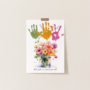 Op de afbeelding: Een witte kaart met drie kleurrijke handafdrukken in groen, oranje en roze. De handafdrukken zijn gelabeld met de namen "Elias", "Lila" en "Marie". Onder de handafdrukken bevindt zich een aquarel van een boeket bloemen in een glazen vaas. De kaart luidt "Mit Liebe von Hand gemacht", wat "Met liefde met de hand gemaakt" betekent.