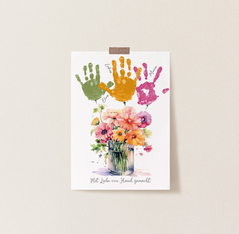 Op de afbeelding: Een witte kaart met drie kleurrijke handafdrukken in groen, oranje en roze. De handafdrukken zijn gelabeld met de namen "Elias", "Lila" en "Marie". Onder de handafdrukken bevindt zich een aquarel van een boeket bloemen in een glazen vaas. De kaart luidt "Mit Liebe von Hand gemacht", wat "Met liefde met de hand gemaakt" betekent.