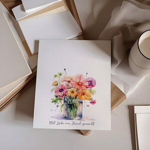 May include: A watercolor painting of a bouquet of pink, orange, and purple flowers in a clear glass vase. The flowers are arranged in a loose, natural style. The painting is signed "Mit Liebe von Hand gemacht" which translates to "Made with love by hand".