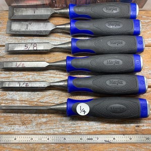 May include: A set of five Marples brand wood chisels with black and blue handles. The chisel blades are labeled with their sizes: 1", 5/8", 1/2", and 1/4". A metal ruler is placed below the chisels, showing inch measurements.