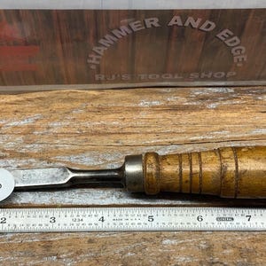 May include: A vintage wood-handled chisel with a metal blade. The handle is light brown with a ribbed design. A sticker with the number 140 is on the blade. A metal ruler is below the chisel.
