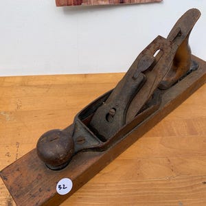 May include: A vintage wooden hand plane with a dark, weathered appearance. The plane features a wooden body, a metal blade, and a rounded knob. The metal components show signs of rust, indicating age and use. The tool is designed for woodworking.