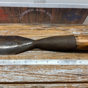 May include: A vintage metal chisel with a wooden handle. The chisel has a wide, flat blade and a dark, aged appearance. A ruler is placed next to the chisel. The sign above the chisel reads "HAMMER AND EDGE - N.J.'S TOOL SHOP."
