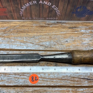 May include: A vintage wood chisel with a metal blade and a wooden handle. The chisel is placed on a wooden surface next to a metal ruler. The handle has a brass end. The tool is approximately 10 inches long.