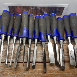 May include: A collection of fifteen metal woodworking chisels arranged in a row, each featuring a black and blue rubberized grip handle and a sharp steel blade. The tools are positioned horizontally with the metal blades pointing downward.