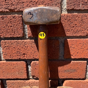 May include: A heavy-duty sledgehammer with a weathered, dark metallic head and a smooth, cylindrical wooden handle. The metal head features the handwritten number 3 in black ink, and a yellow circular sticker with the number 93 is attached.