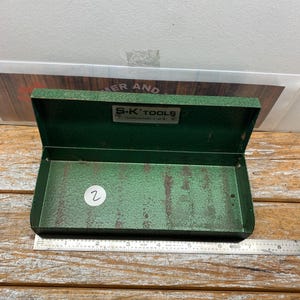May include: Open green metal tool box with the text "S-K TOOLS" on the lid. The box is open, revealing the interior. A metal ruler is next to the box. The box is on a wooden surface.