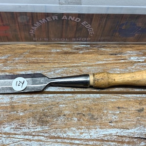 May include: A metal chisel with a wooden handle. The chisel has a wide, flat blade and a dark metal body. The handle is light-colored wood with a darker end. A sticker with the number 124 is on the blade.
