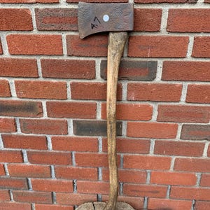 May include: An axe with a wooden handle and a metal head, resting on a tree stump. The axe head has the number "A7" written on it. The background is a red brick wall.