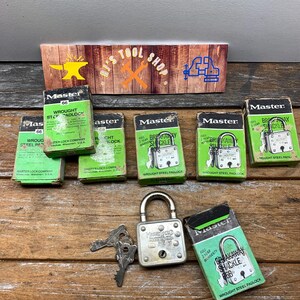 May include: A collection of six vintage Master Lock padlocks in green and white boxes. The boxes feature the Master Lock logo and the text "Wrought Steel Padlock". The padlocks are silver and have a break-away shackle. The boxes are on a wooden surface.