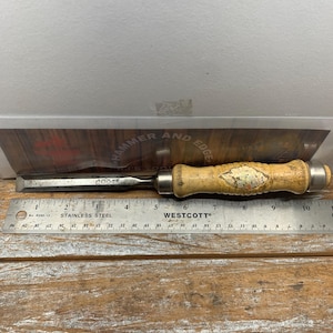 May include: A wood-handled chisel with a metal blade, lying on a wooden surface next to a stainless steel ruler. The chisel has a light-colored wooden handle with a decorative design. The ruler is marked with both inches and centimeters.