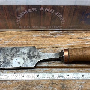 May include: A vintage wood chisel with a worn, dark metal blade and a wooden handle. The blade is approximately 11 inches long. The handle is a light brown color with a metal end cap. The tool is resting on a wooden surface.