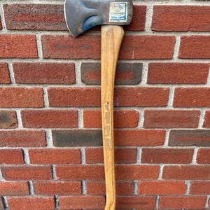 May include: A Collins Commander axe with a curved wooden handle and a dark metal head. The head features a blue-tinted finish and a rectangular label with the text Commander Collins Axe. The handle has printed safety text along its length.