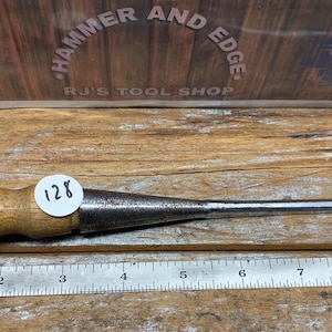 May include: A Craftsman wood chisel with a wooden handle and a metal blade. The chisel has a round wooden handle and a metal blade. A sticker with the number 128 is on the handle. A metal ruler is below the chisel.