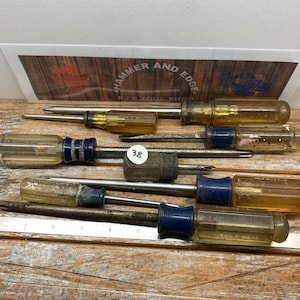 May include: A collection of screwdrivers with clear and blue handles, and metal shafts. The tools are arranged on a wooden surface. The image also shows a sign that reads "HAMMER AND EDGE RU'S TOOL SHOP".