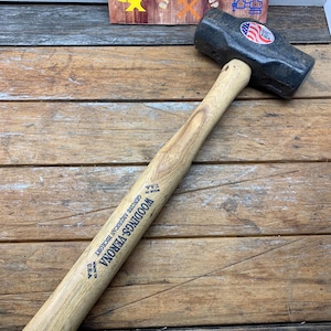 May include: A black metal sledgehammer with a wooden handle. The handle is labeled "Woodings-Verona" and "Genuine American Hickory" with "Made in U.S.A."