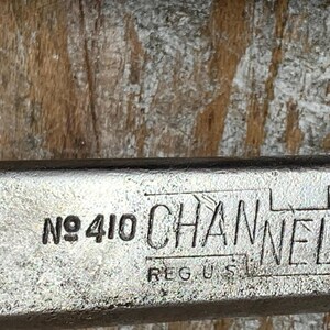 K&ouml;nnte beinhalten: Nahaufnahme eines Metallwerkzeugs mit dem Text "No 410 CHANNEL LOCK REG. U.S. PAT." Das Werkzeug hat eine silberne Oberfl&auml;che und befindet sich vor einem Holzgrund. Der Text ist schwarz.