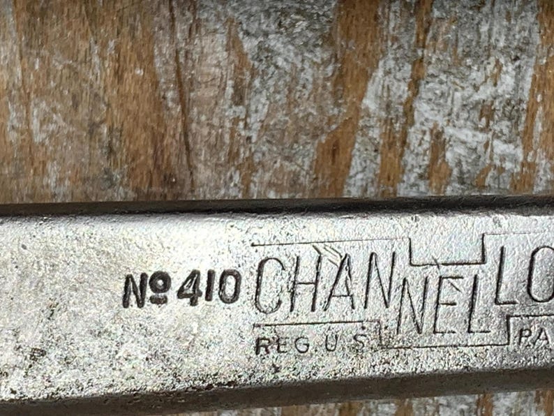 K&ouml;nnte beinhalten: Nahaufnahme eines Metallwerkzeugs mit dem Text "No 410 CHANNEL LOCK REG. U.S. PAT." Das Werkzeug hat eine silberne Oberfl&auml;che und befindet sich vor einem Holzgrund. Der Text ist schwarz.