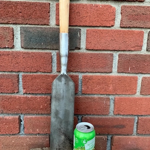 May include: A large, weathered metal garden tool with a wooden handle. The tool is next to a green can of lemon-lime soda. The background is a red brick wall. The tool appears to be a type of spade or edging tool.