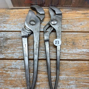 K&ouml;nnte beinhalten: Zwei silberfarbene Kanalzangen aus Metall mit strukturierten Griffen und verstellbaren Backen. Auf den Zangen steht "420 CHANNEL LOCK" und "CHAMPION DE ARMENT". Ein Preisschild mit der Nummer "83" ist an einer der Zangen befestigt. Der Hintergrund enth&auml;lt den Text "HAMMER AND EDGE" und "RJ'S TOOL SHOP".
