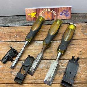 May include: Three Stanley chisels with yellow and black handles and three black plastic chisel holders. The chisels are on a wooden surface.