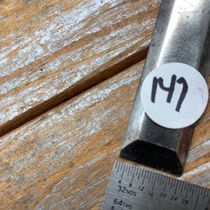 May include: A metal chisel with a beveled edge, next to a metal ruler. The chisel has a sticker with the number 147 on it. The ruler shows measurements in inches and fractions.